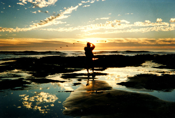 A man stands on rocks in tidepools, taking a photo of pelicans flying past.